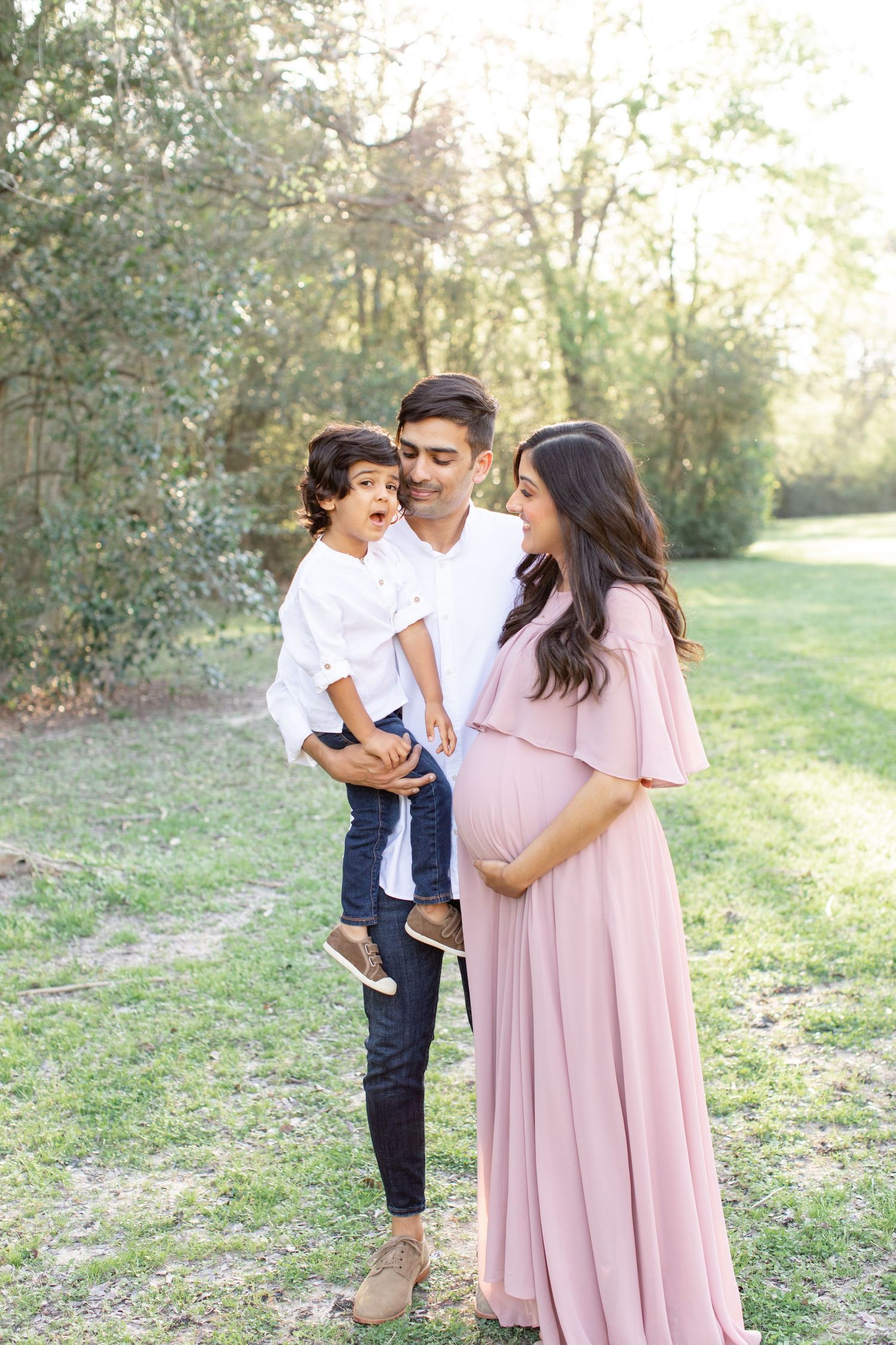 A family shares a joyful moment together outdoors, with the woman wearing a pink maternity gown.