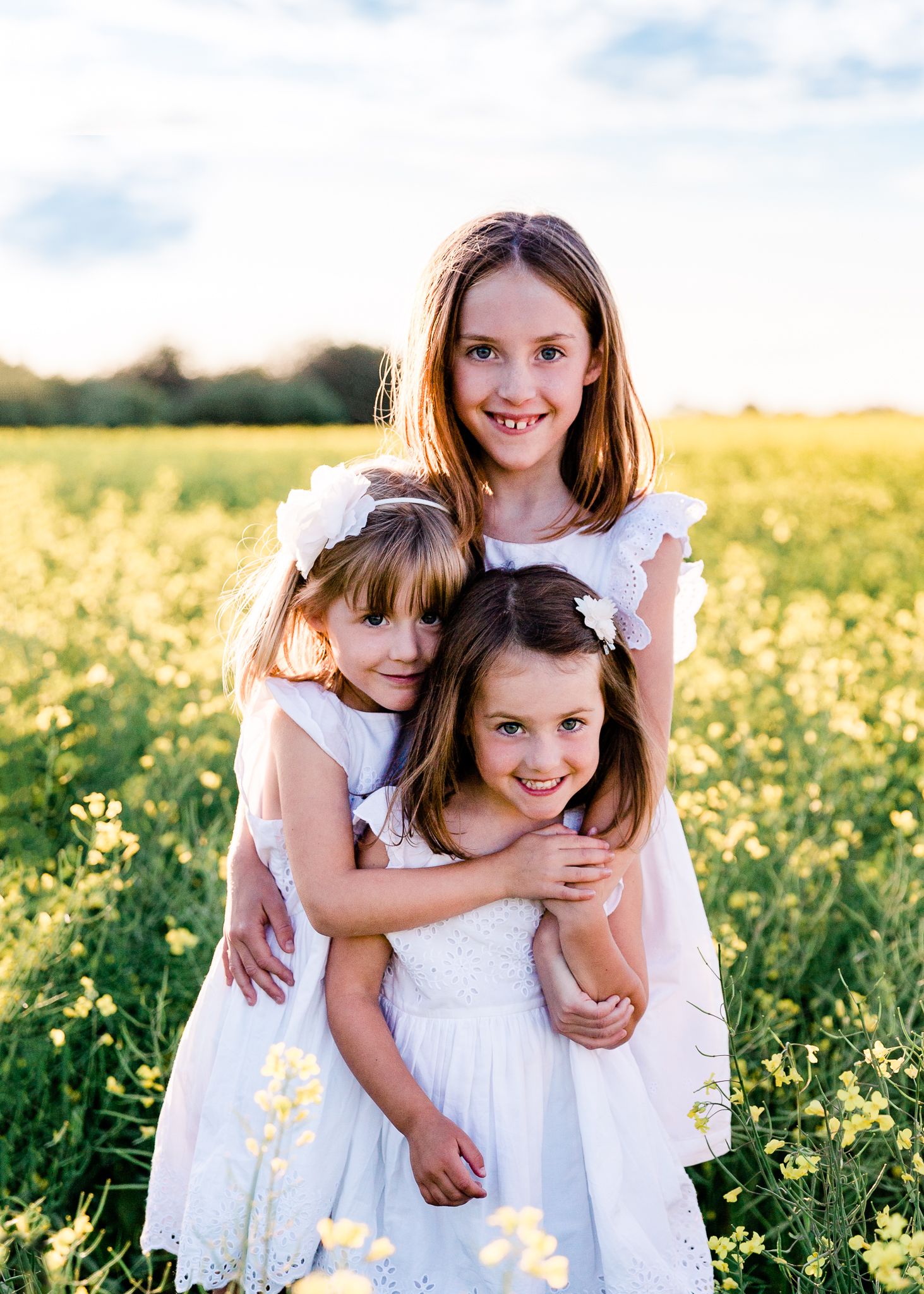 Three sisters in white dresses hugging in a vibrant yellow flower field.