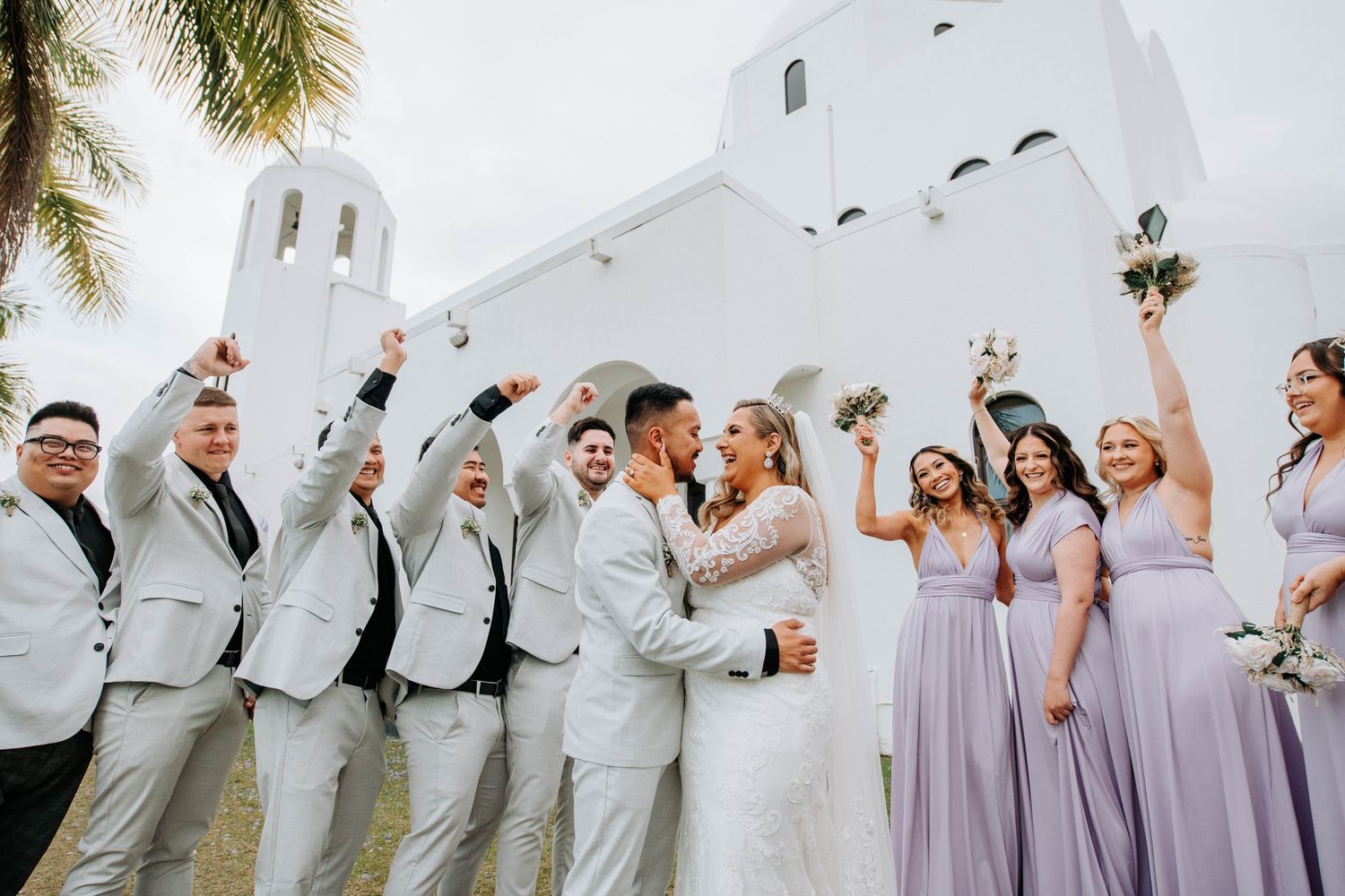 Wedding party celebrates outside white Spanish-style church in coordinated attire.