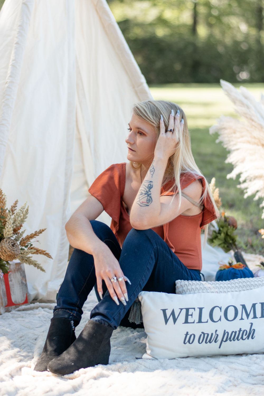 Person in rust-colored top sitting by teepee with welcome sign and dried pampas grass decorations.