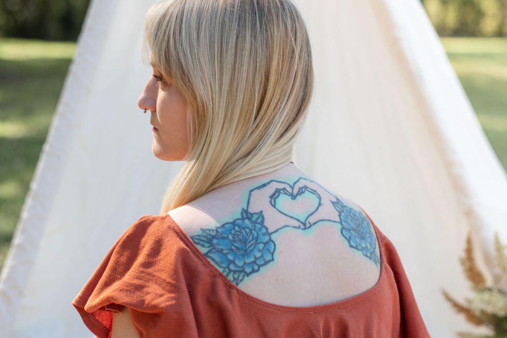 A close-up of a blue heart tattoo design on the upper back visible through a rust-colored top.
