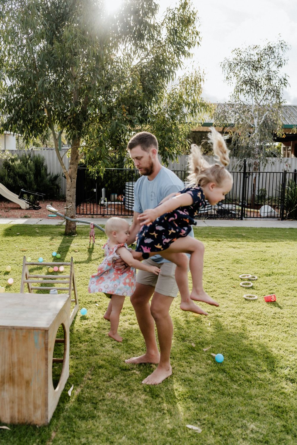 Two young girls jumping off play equipment in their backyard, with their Dad watching