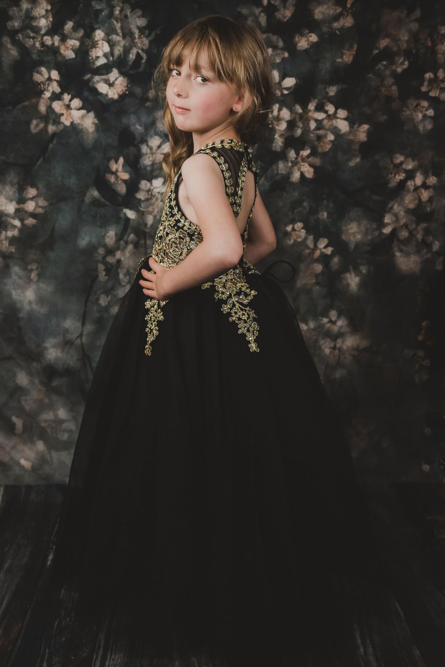 Young girl in black and gold tulle ball gown poses against floral backdrop in studio.