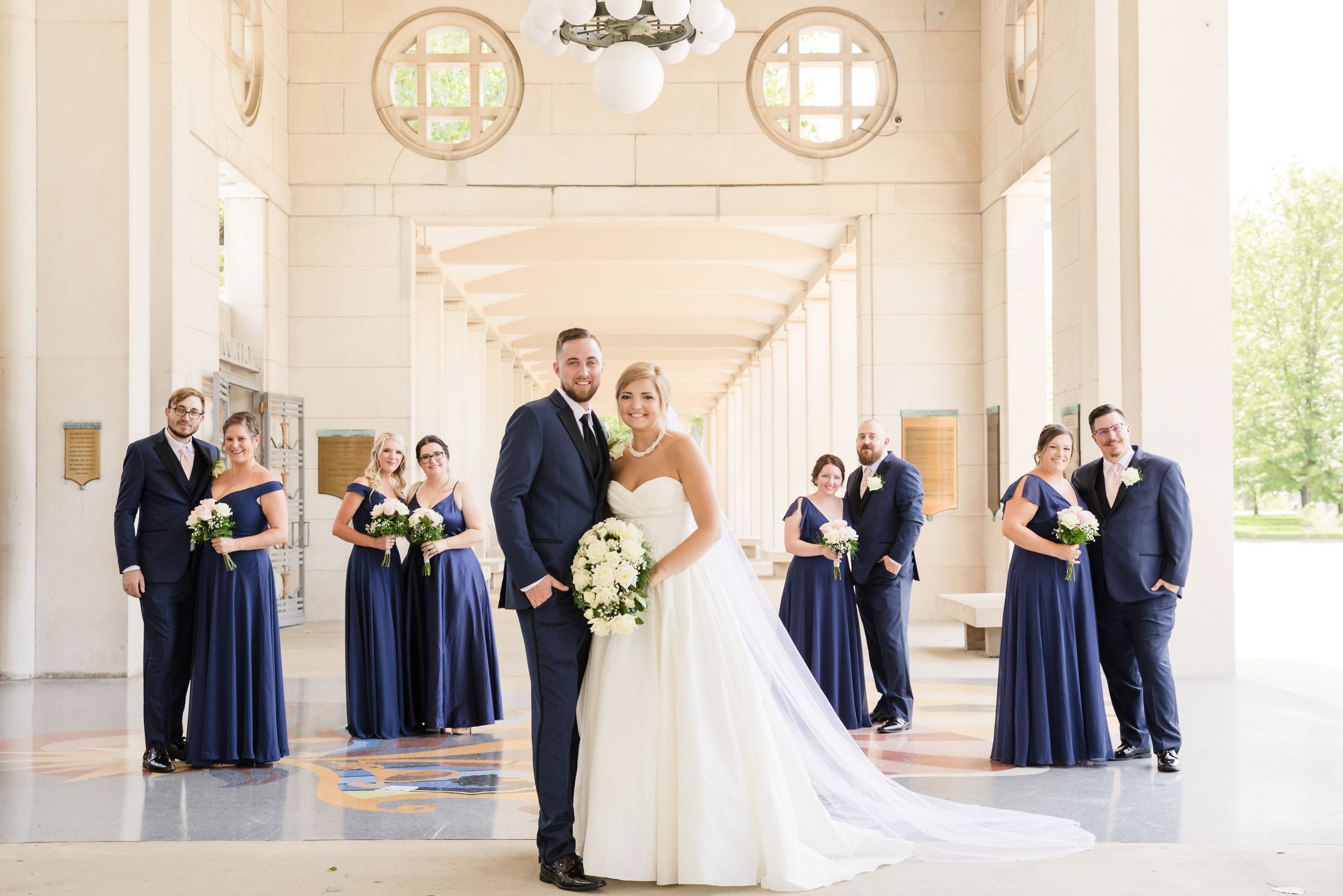 A wedding party in navy blue attends to the couple in an elegant white columned hallway.