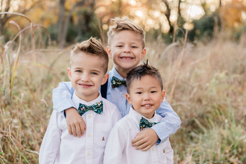 Three young boys in matching white shirts and bowties pose together in tall grass.