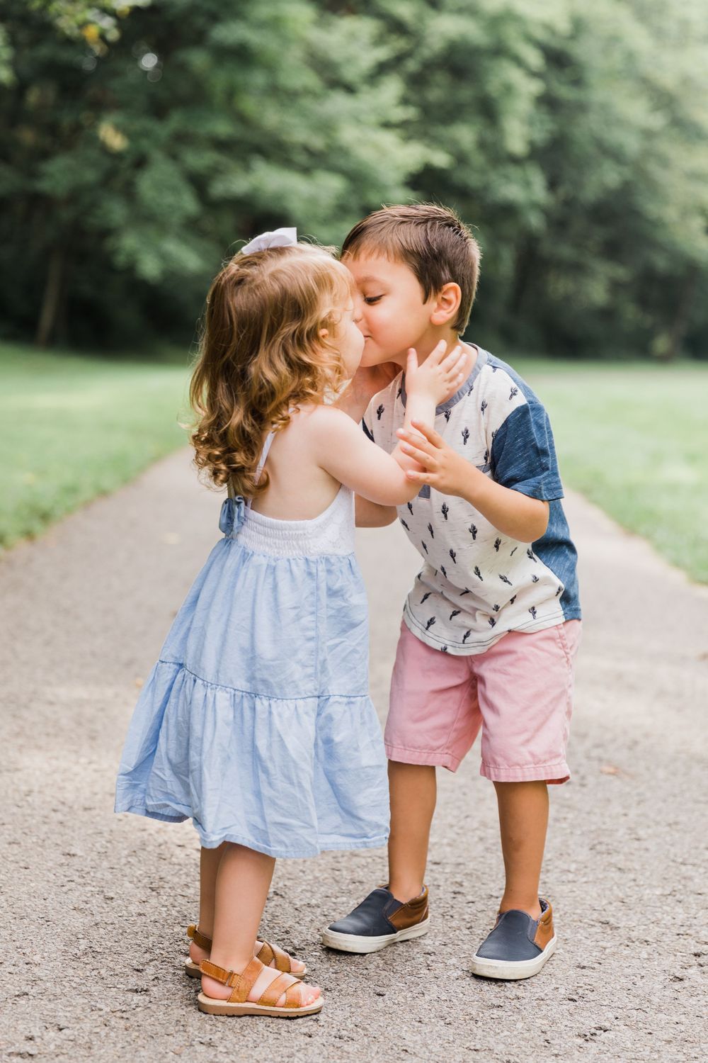 Two children sharing a sweet kiss on a tree-lined path while wearing summer outfits.