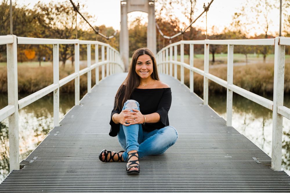 Portrait of woman in black top sitting cross-legged on white bridge at sunset.