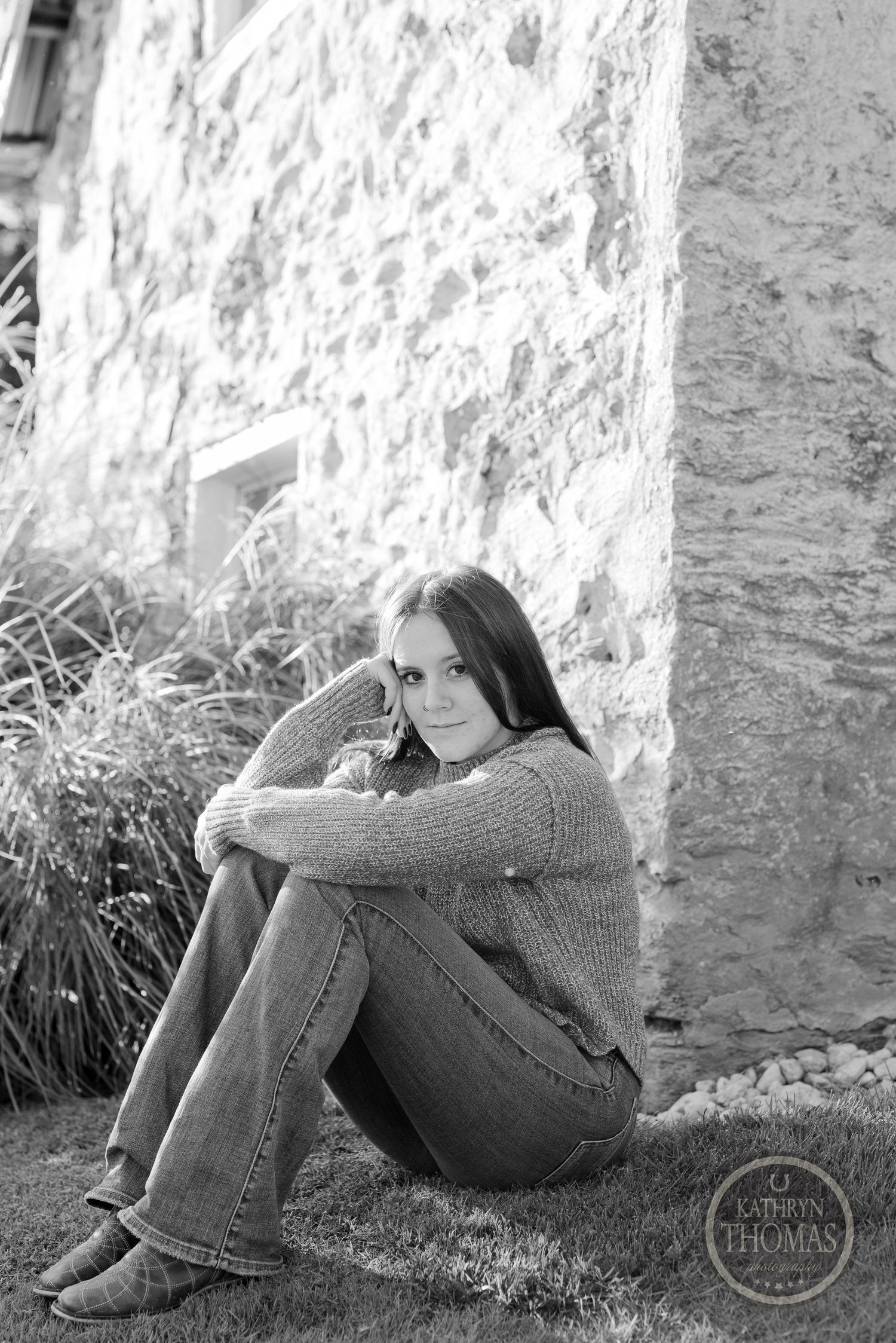 Black and white portrait of someone sitting against stone wall wearing sweater and jeans.
