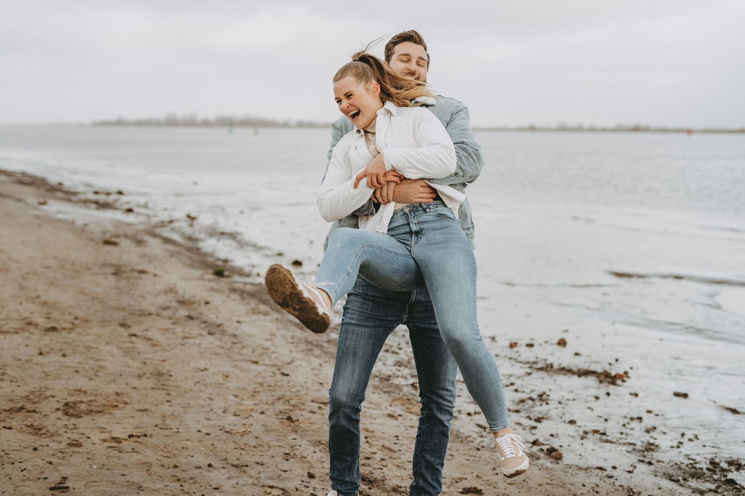 A playful couple embracing on a beach with cloudy skies in the background.