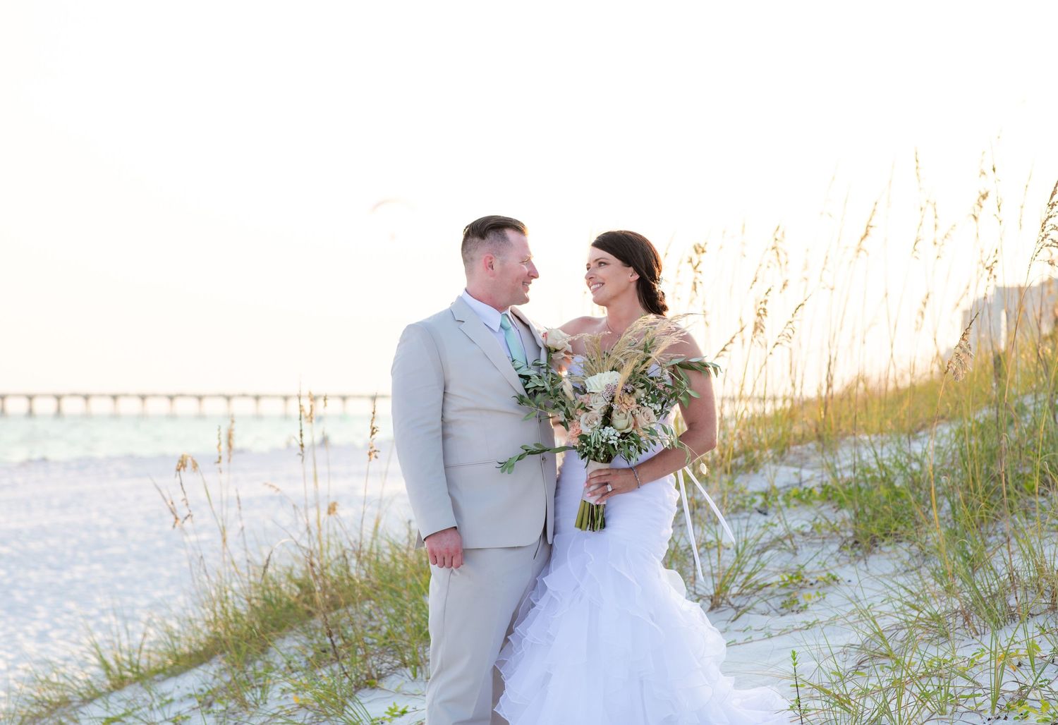 A beach wedding couple poses together on sand dunes with a pier visible in the background.