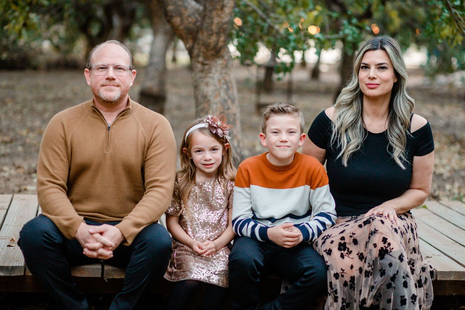 Family of four sitting on a wooden bench outdoors, parents flanking two young children in a wooded park setting.