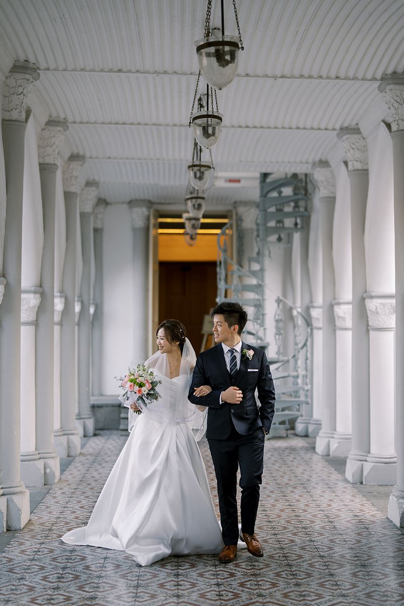 Wedding couple walks through elegant white colonnade hallway with vintage lighting.