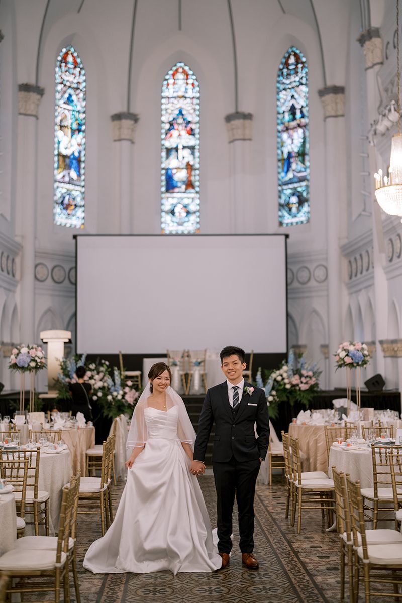 Wedding couple poses in church reception hall with stained glass windows and elegant table settings.