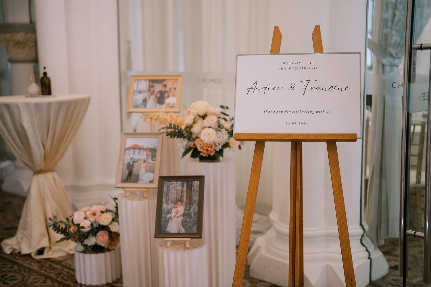 Elegant wedding decor display with white pedestals, floral arrangements, and framed photos against a light backdrop.
