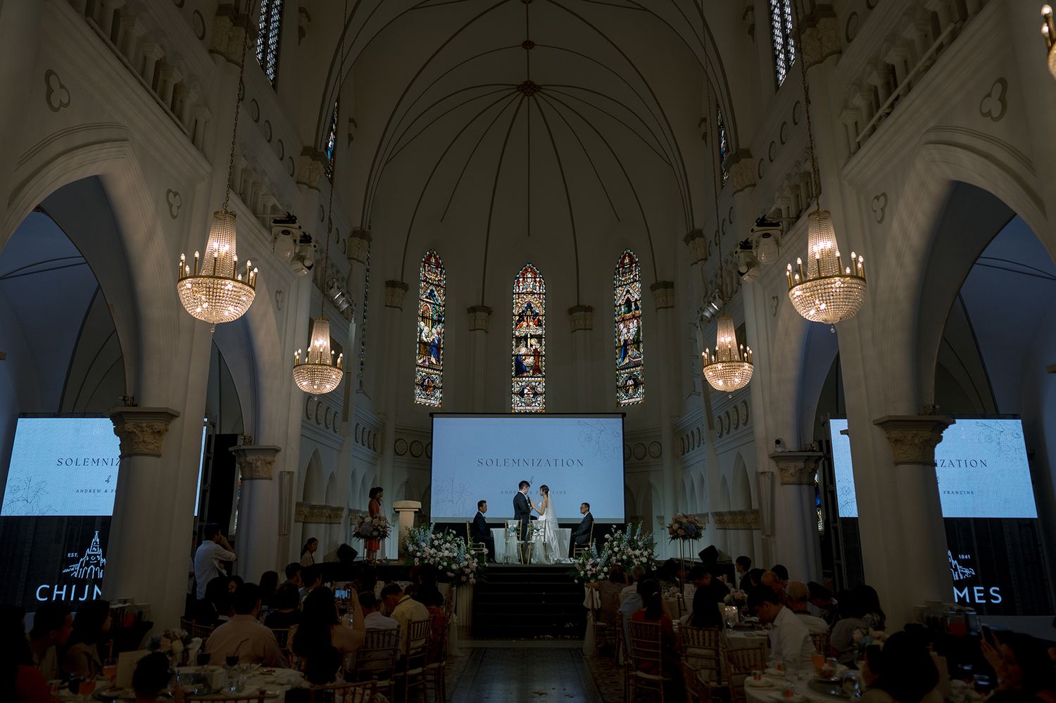 Elegant church interior with vaulted ceilings, stained glass windows, and hanging chandeliers during a wedding ceremony.