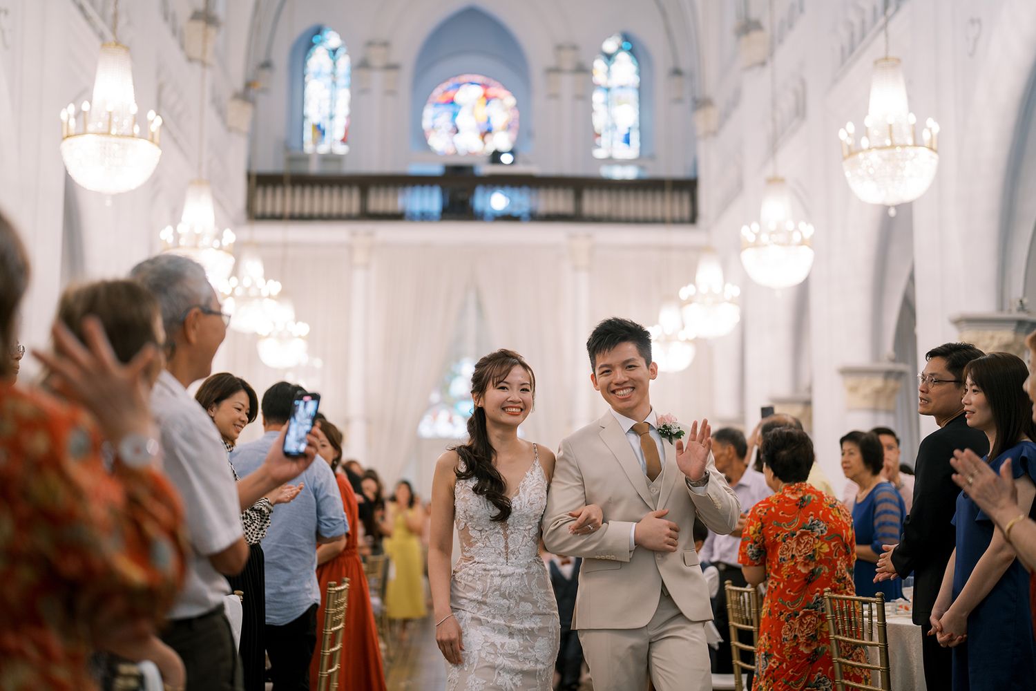 Wedding guests celebrate in an elegant white church interior with chandeliers and stained glass windows.
