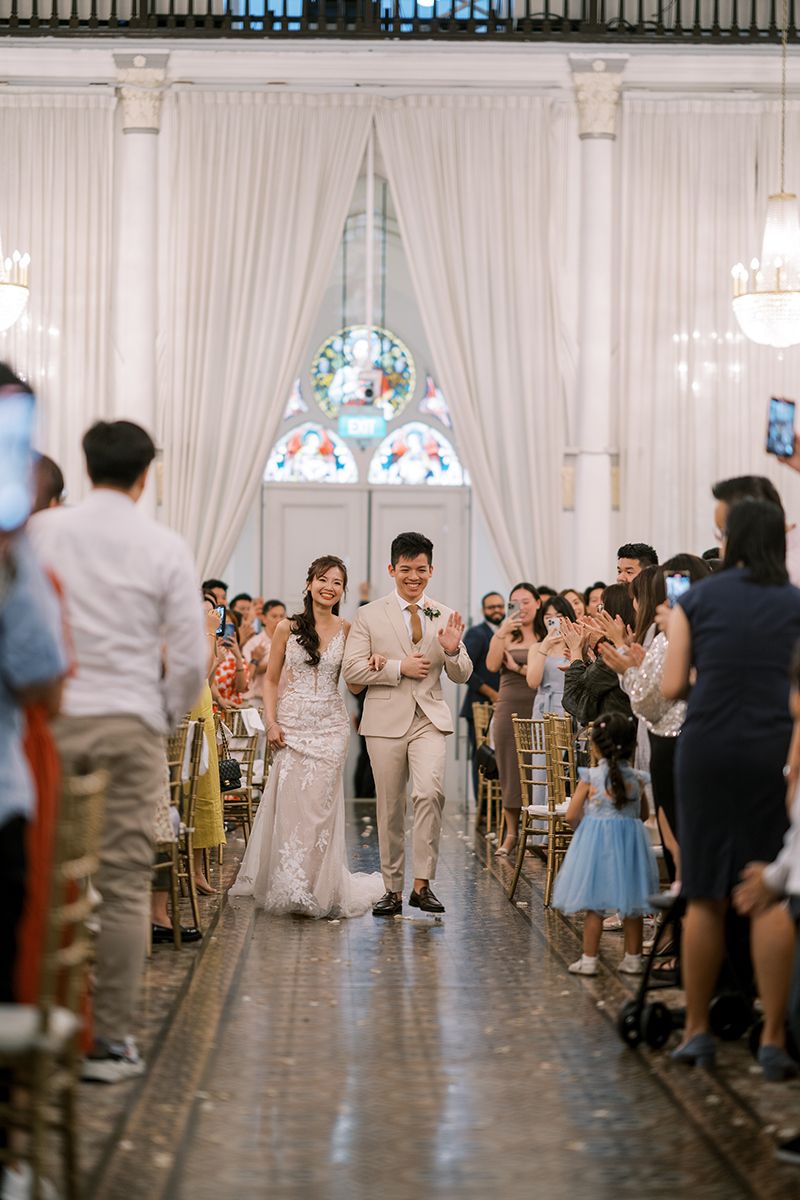 Wedding guests stand and celebrate in an elegant white reception hall with draped walls.
