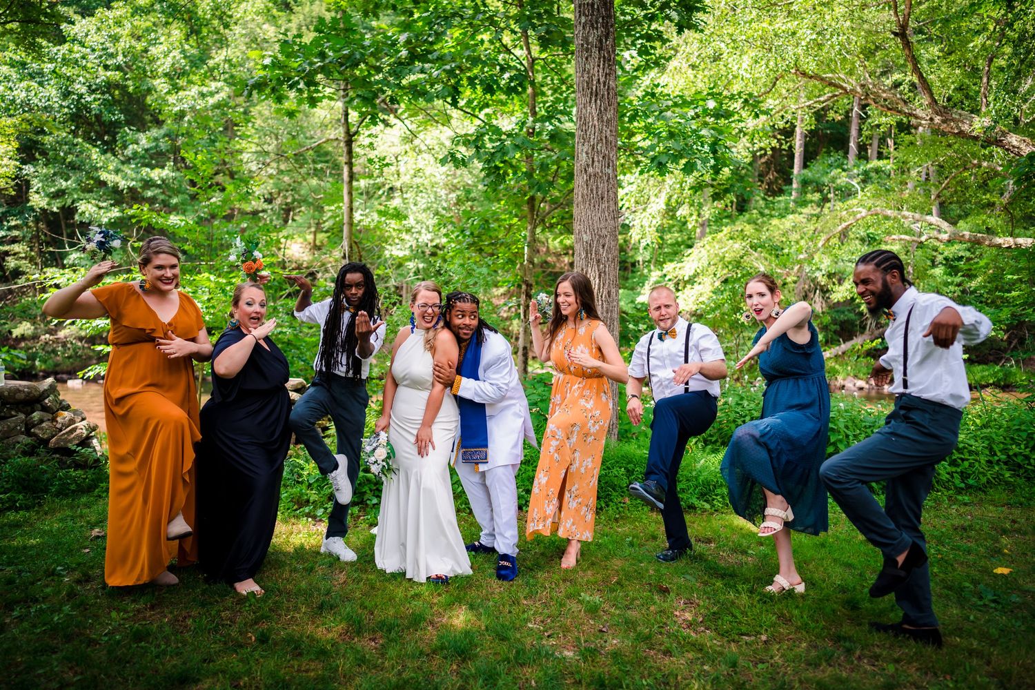 Fun wedding party group shot with everyone striking dance poses in wooded setting.