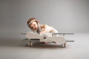 Toddler sitter portrait, child on wooden aeroplane photo prop wear pilots hat, back drop grey prop grey & white.