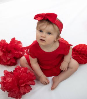 child valentine red photo shoot. red bow headband, red bodysuit,  white back lit   background red tissue pom poms