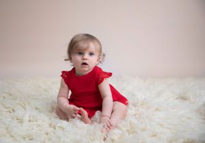 toddler sitter session, baby wearing  red bodysuit dress sitting on cream flokati rug against cream background