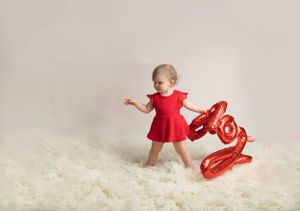 kids valentine photo shoot, child wearing red bodysuit dress holding red foil inflatable LOVE balloon on a cream flokati