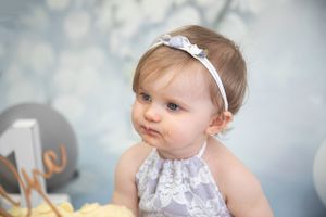 child portrait 1 year cake smash, blue floral backdrop, child wearing purple lace bodysuit & matching head band/tieback