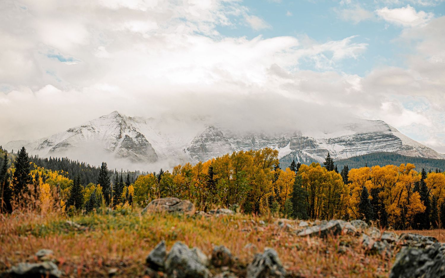 Panoramic mountain landscape with snow-capped peaks and golden autumn foliage against cloudy skies.