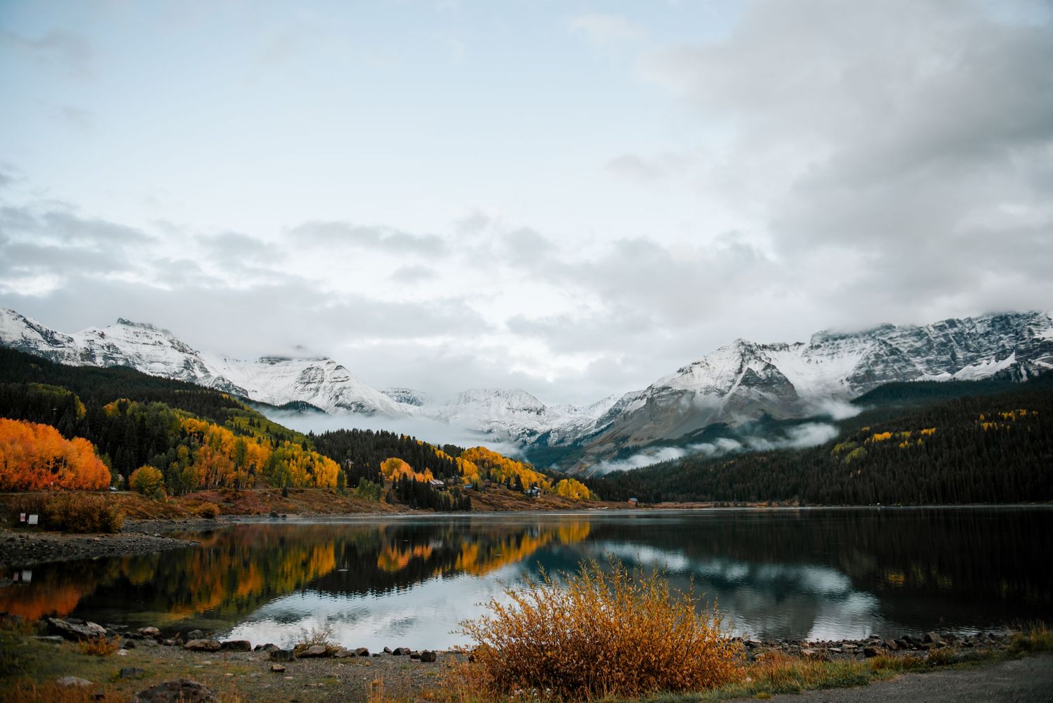 Snow-capped mountains reflect in a calm lake surrounded by golden fall foliage.