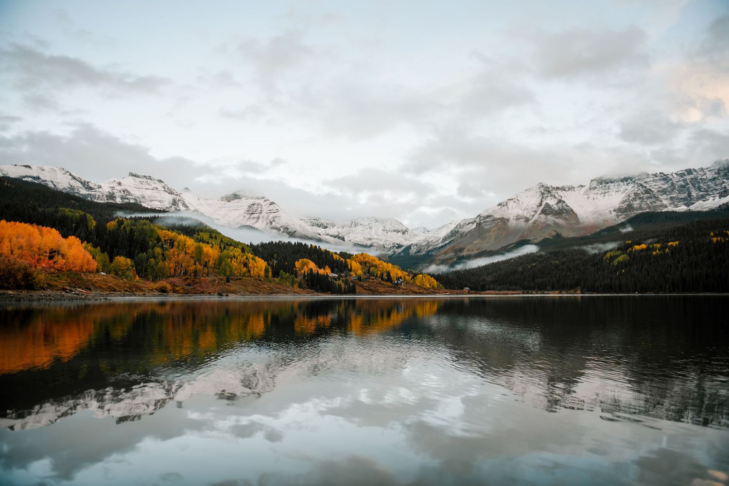 Panoramic view of a mountain lake with snow-capped peaks and fall colors reflecting in still water.
