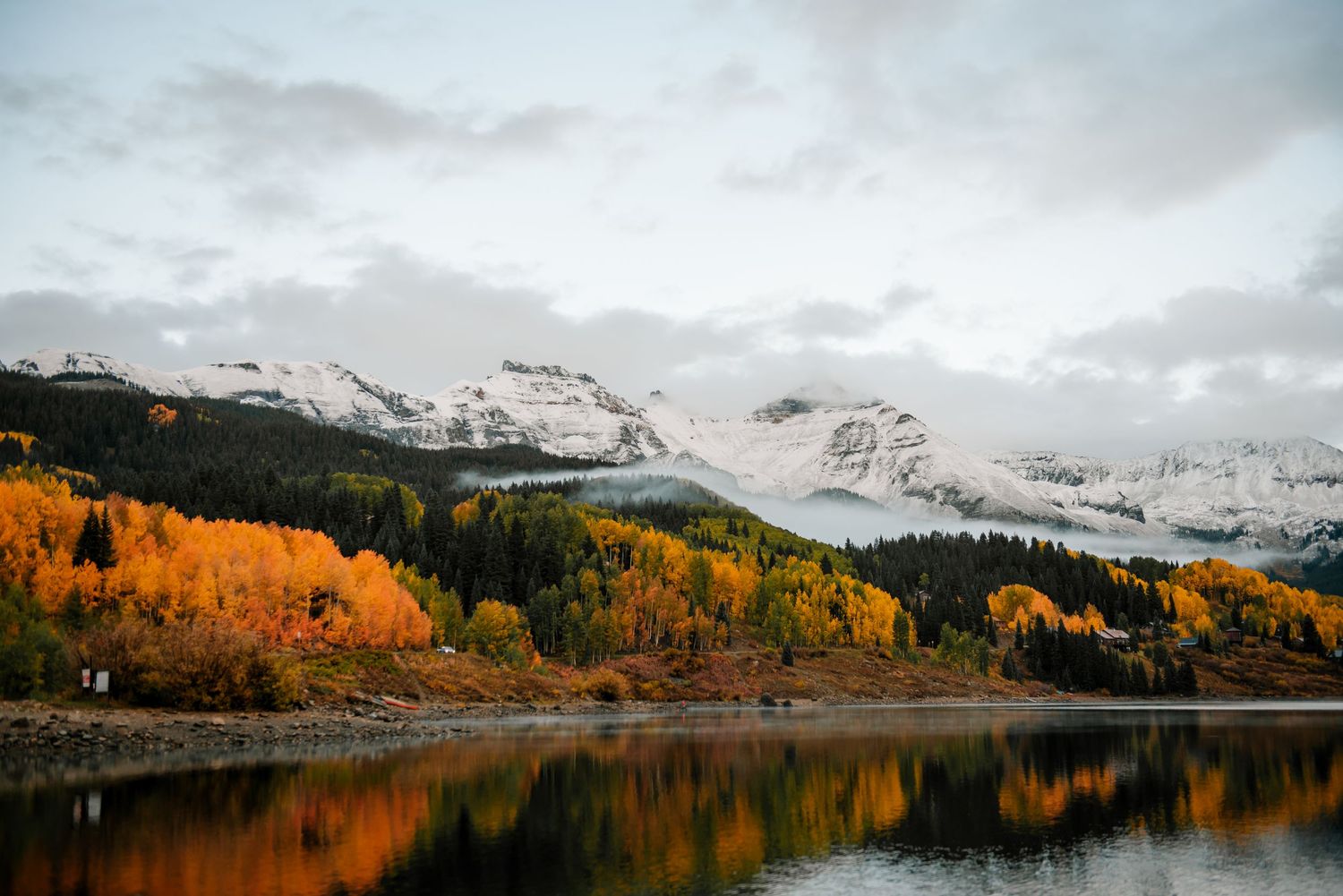Wide mountain landscape with snow-covered peaks and golden autumn trees reflecting in a serene lake.
