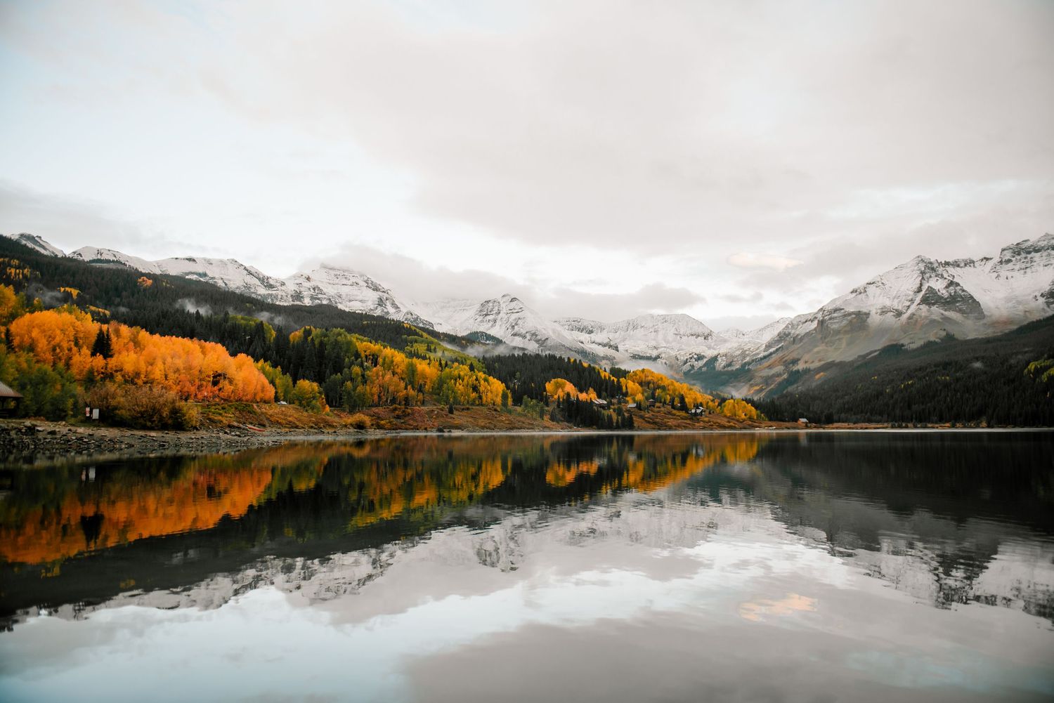 Panoramic view of a mountain lake surrounded by snow-covered peaks and golden autumn trees.