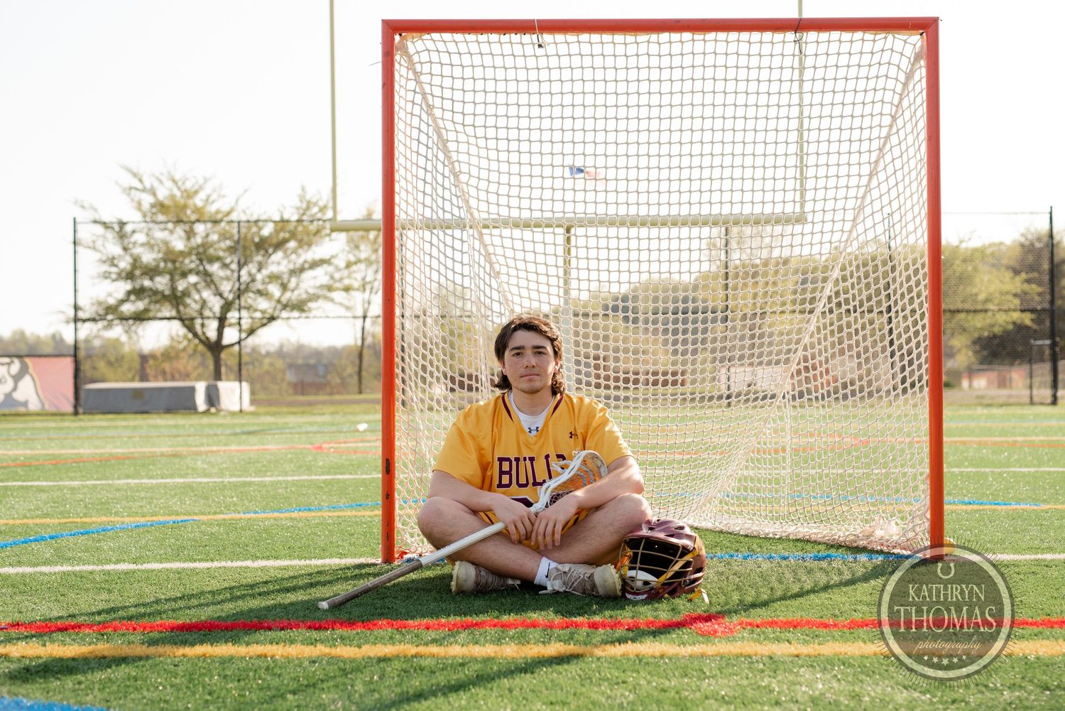 Lacrosse player in yellow Bulls jersey sitting in front of goal net on sports field.
