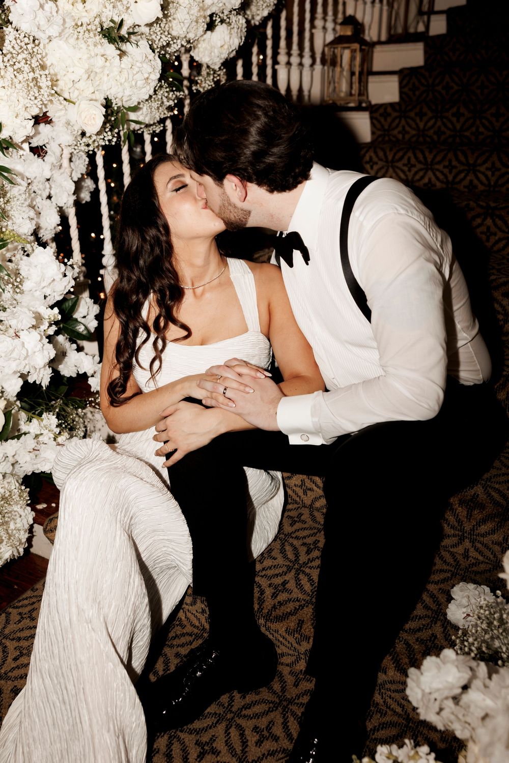 Bride and groom share passionate kiss seated among white floral arrangements on ornate staircase.