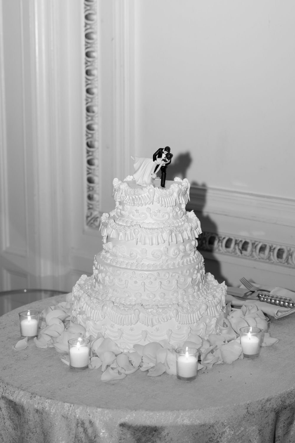 Black and white photo of tiered wedding cake with bride and groom topper surrounded by candles.