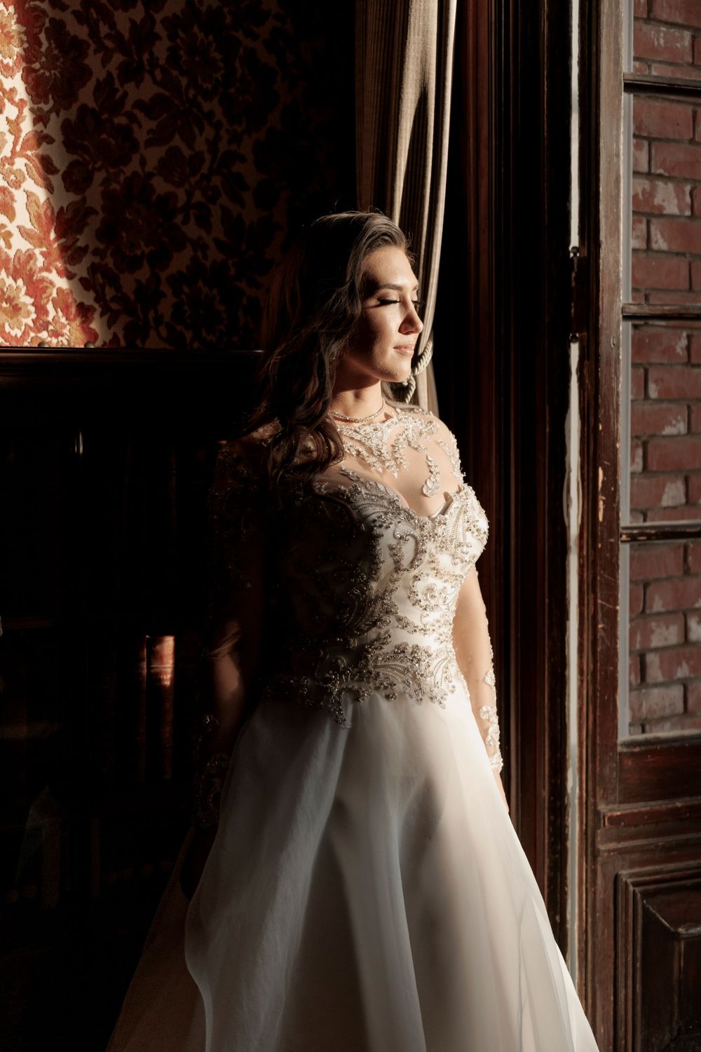 Bride in ornate beaded lace wedding gown stands by tall wooden door in dramatic moody lighting.