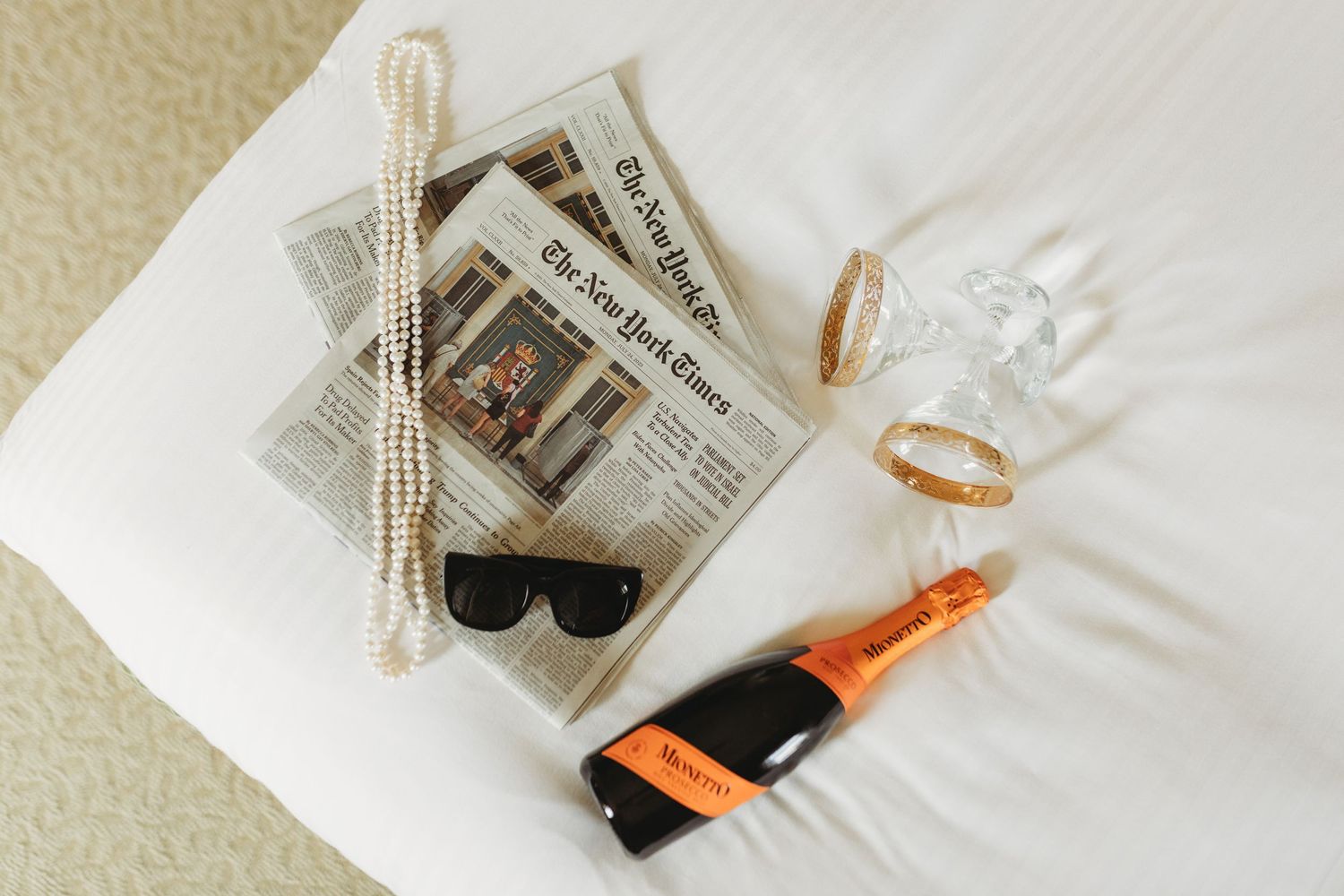 Wedding accessories including champagne, sunglasses, pearls and newspaper laid out on white bedding.