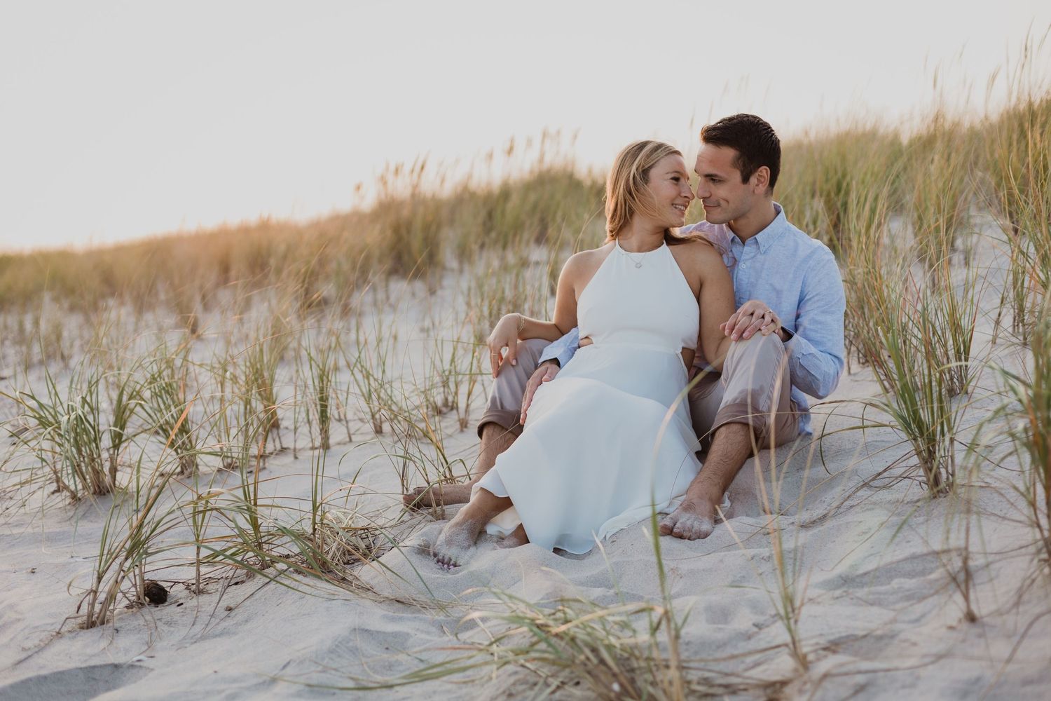 A romantic couple sits together on sandy beach dunes at sunset.