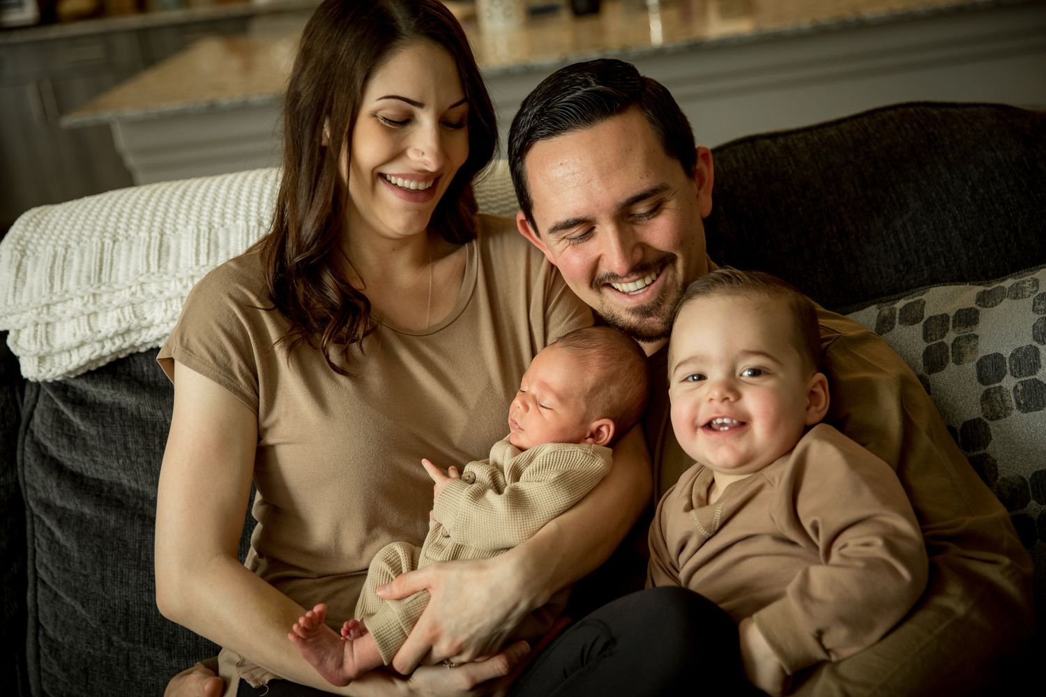 Family relaxes together on couch with newborn and toddler in matching neutral tones.