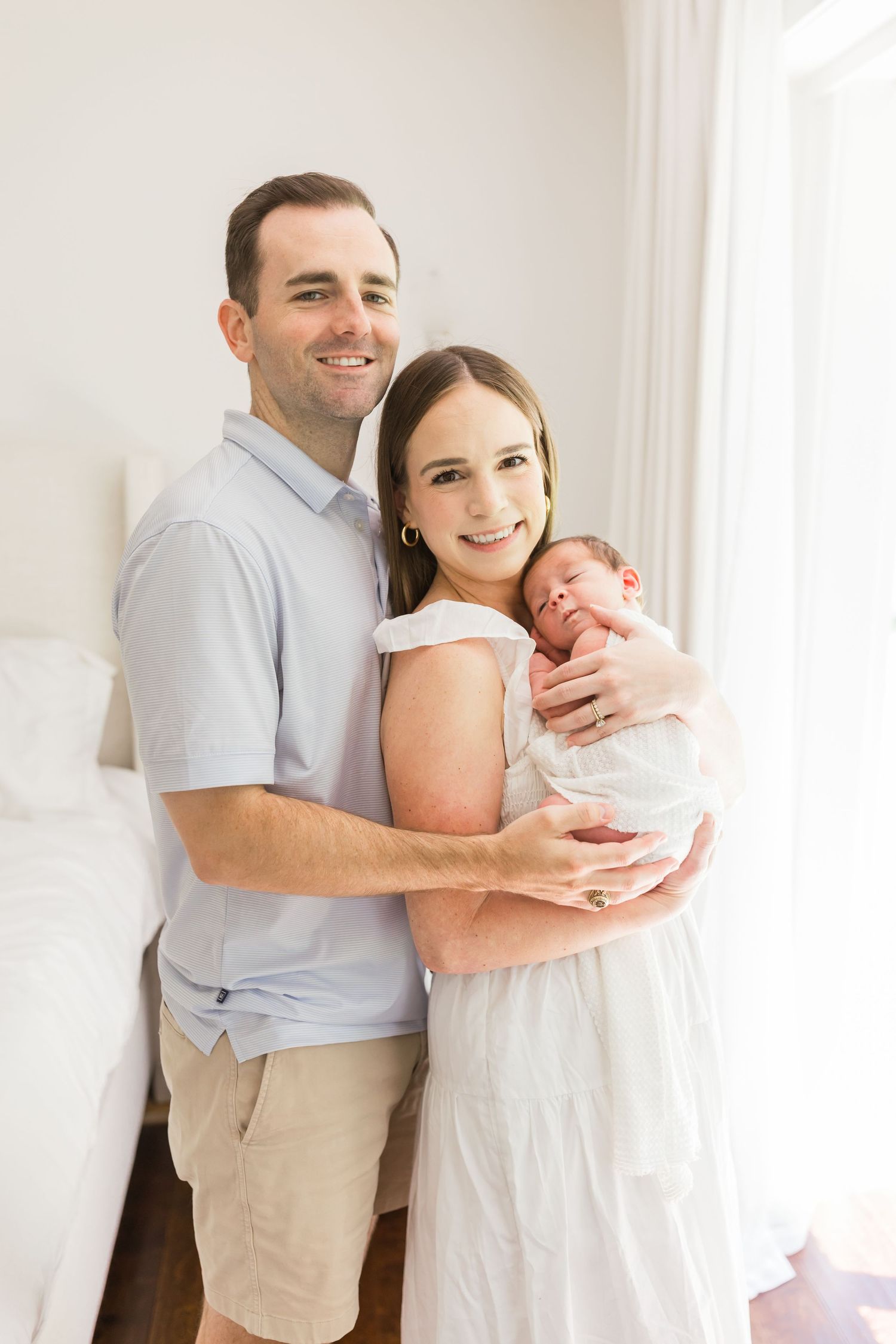 A happy couple holds their newborn baby together while standing against a white wall.