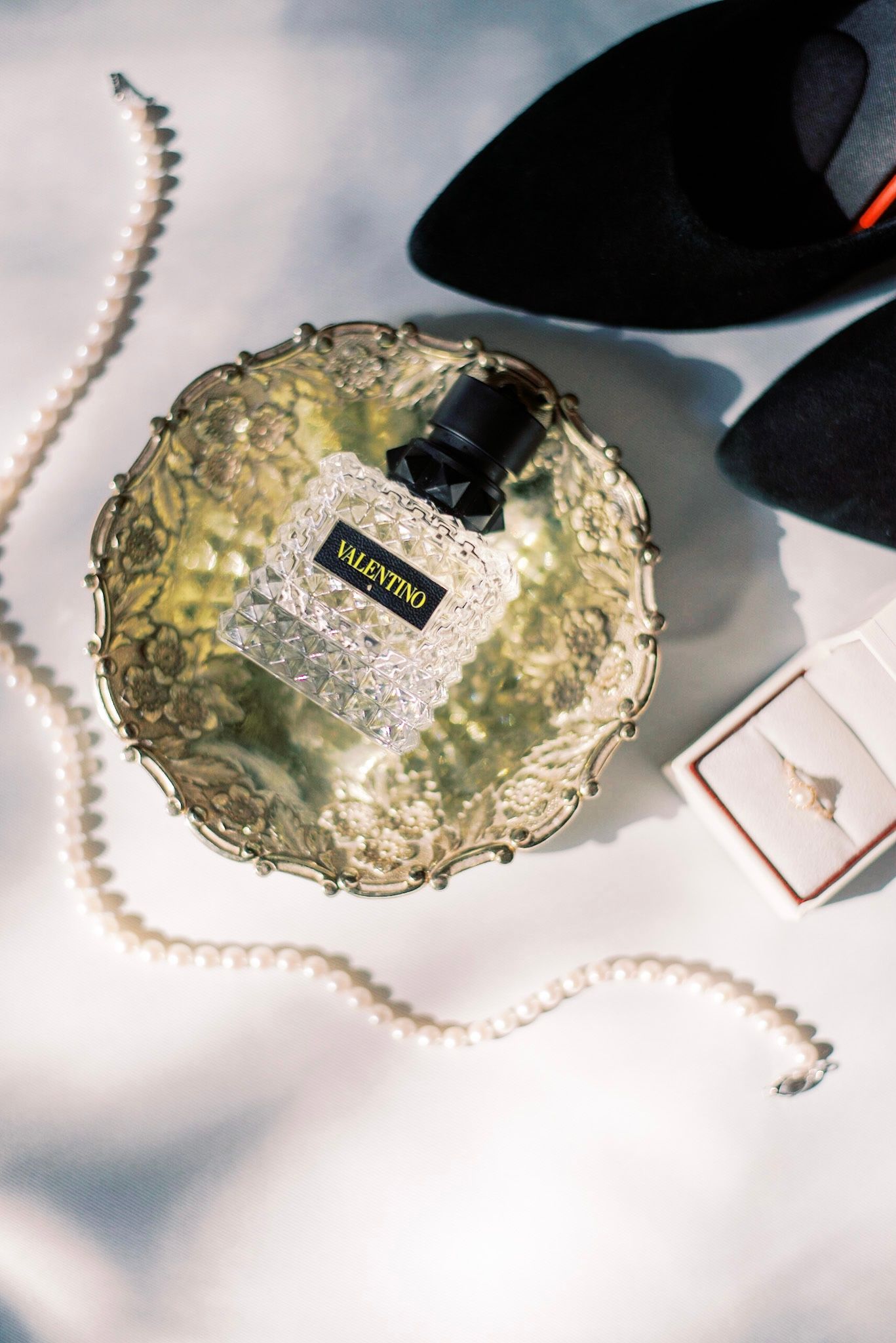 Perfume bottle on ornate silver tray with pearl necklace and black accessories.