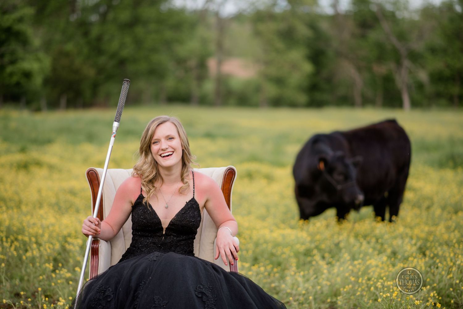 A figure in a black formal dress sits in a chair in a field of yellow flowers with a cow grazing in the background.