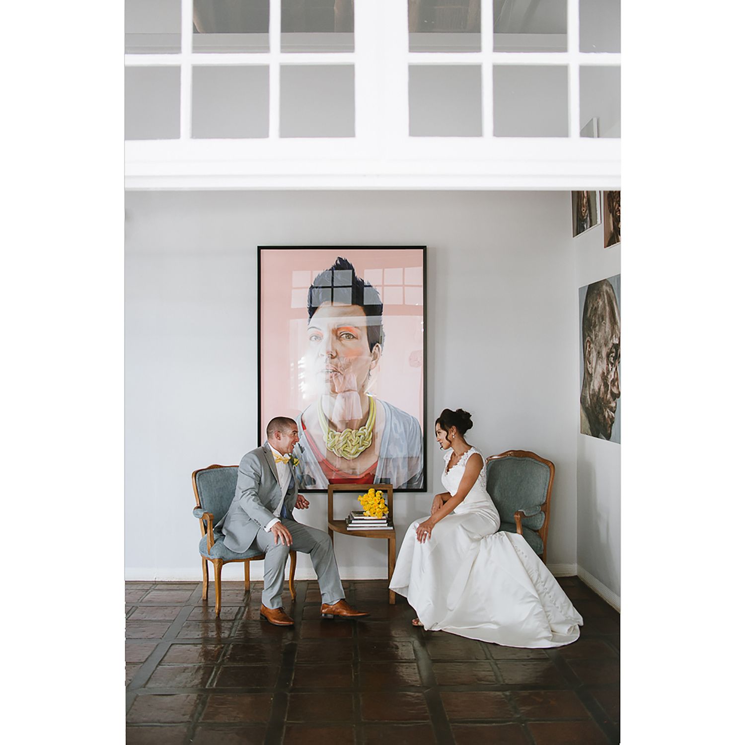 Elegant wedding portrait with couple seated beneath modern art in minimalist white space.