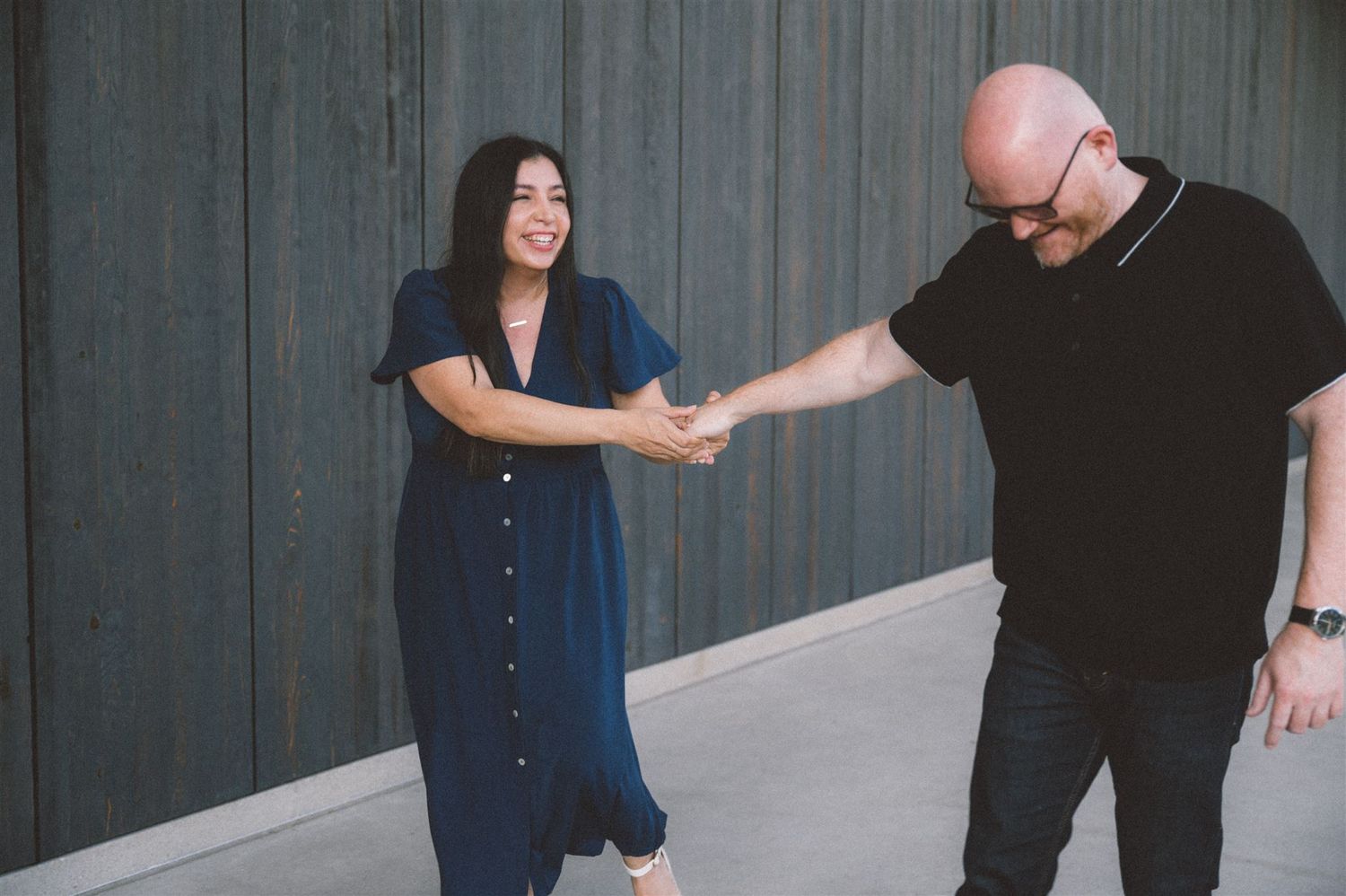 A playful couple in blue dress and black attire dance together on a concrete walkway.