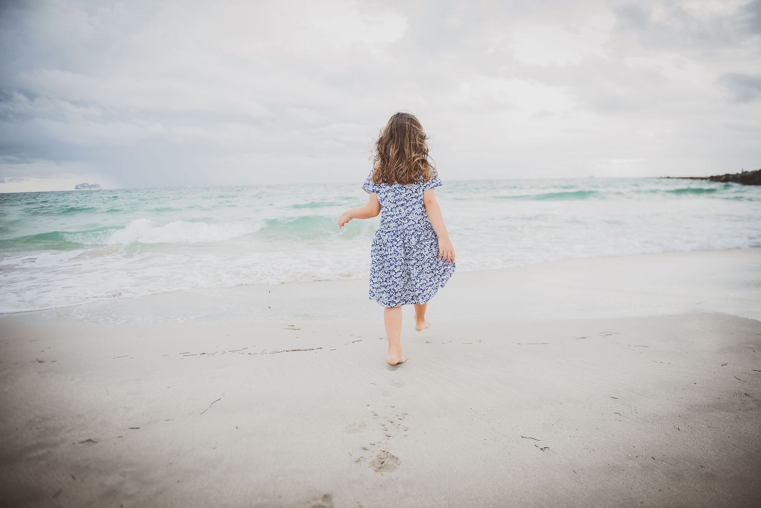 Small girl in a blue polka dot dress runs along a misty beach leaving footprints in the sand.