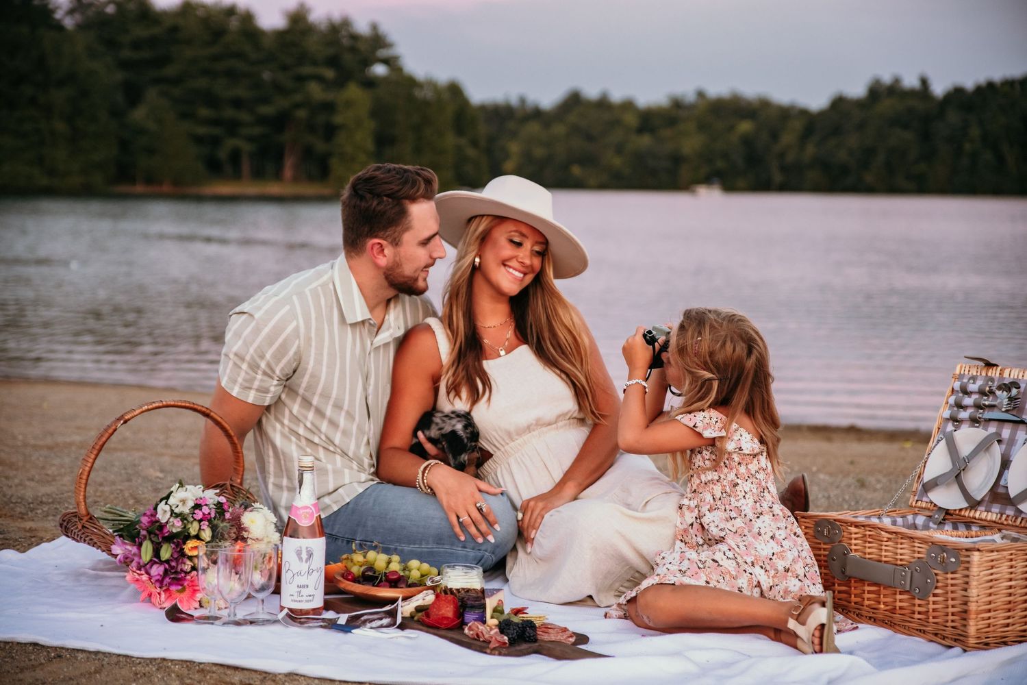 Family enjoying a picnic by the lake at sunset with food basket and flowers.