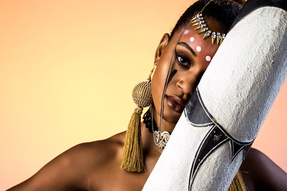 Fashion portrait featuring ornate gold jewelry and dramatic makeup against a peach-colored background.