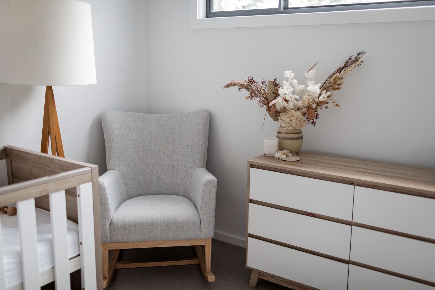 Modern nursery corner with gray armchair, white dresser and dried floral arrangement.