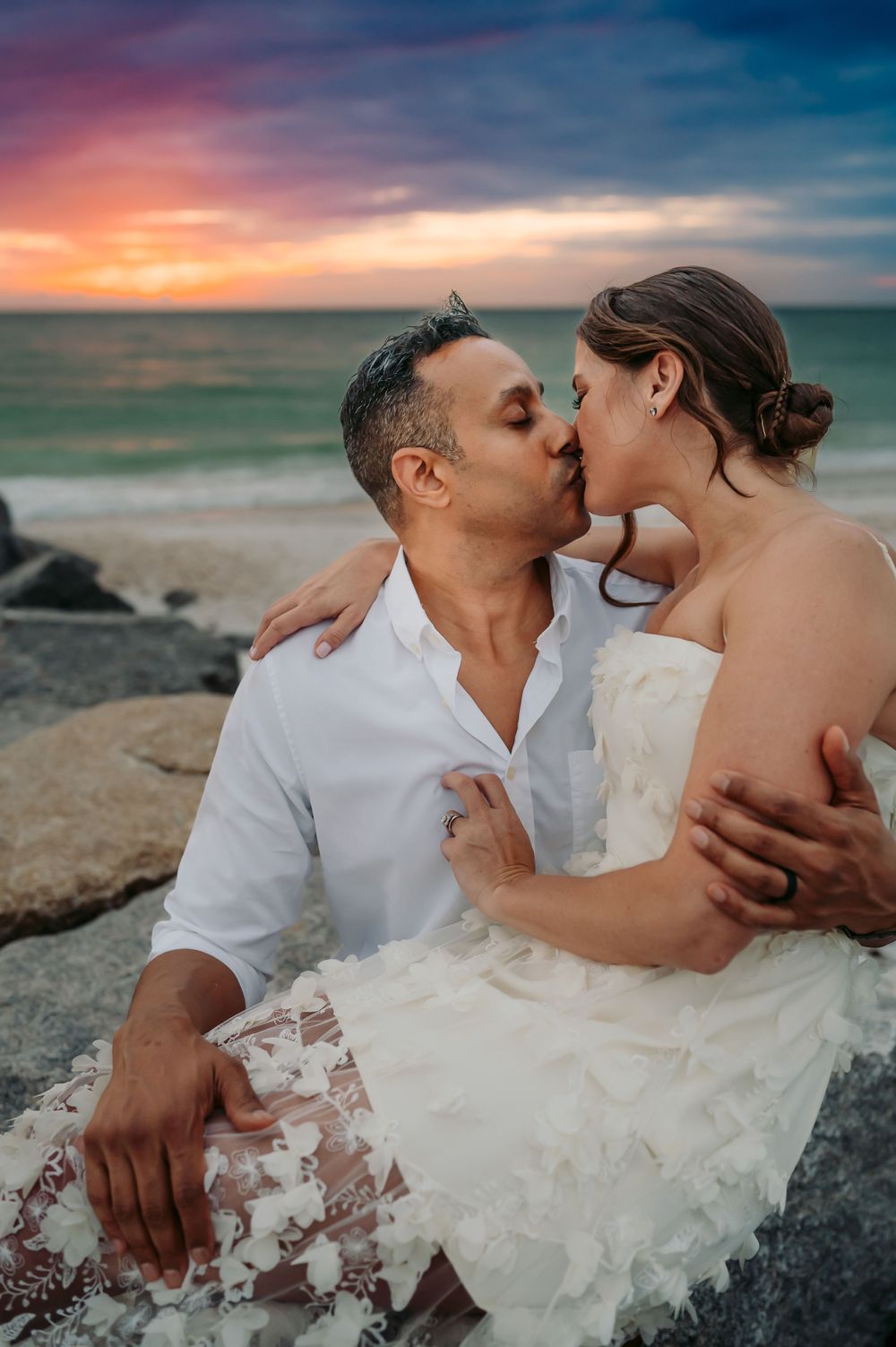 Man and woman at the beach sitting on large rocks and kissing in a passionate embrace.