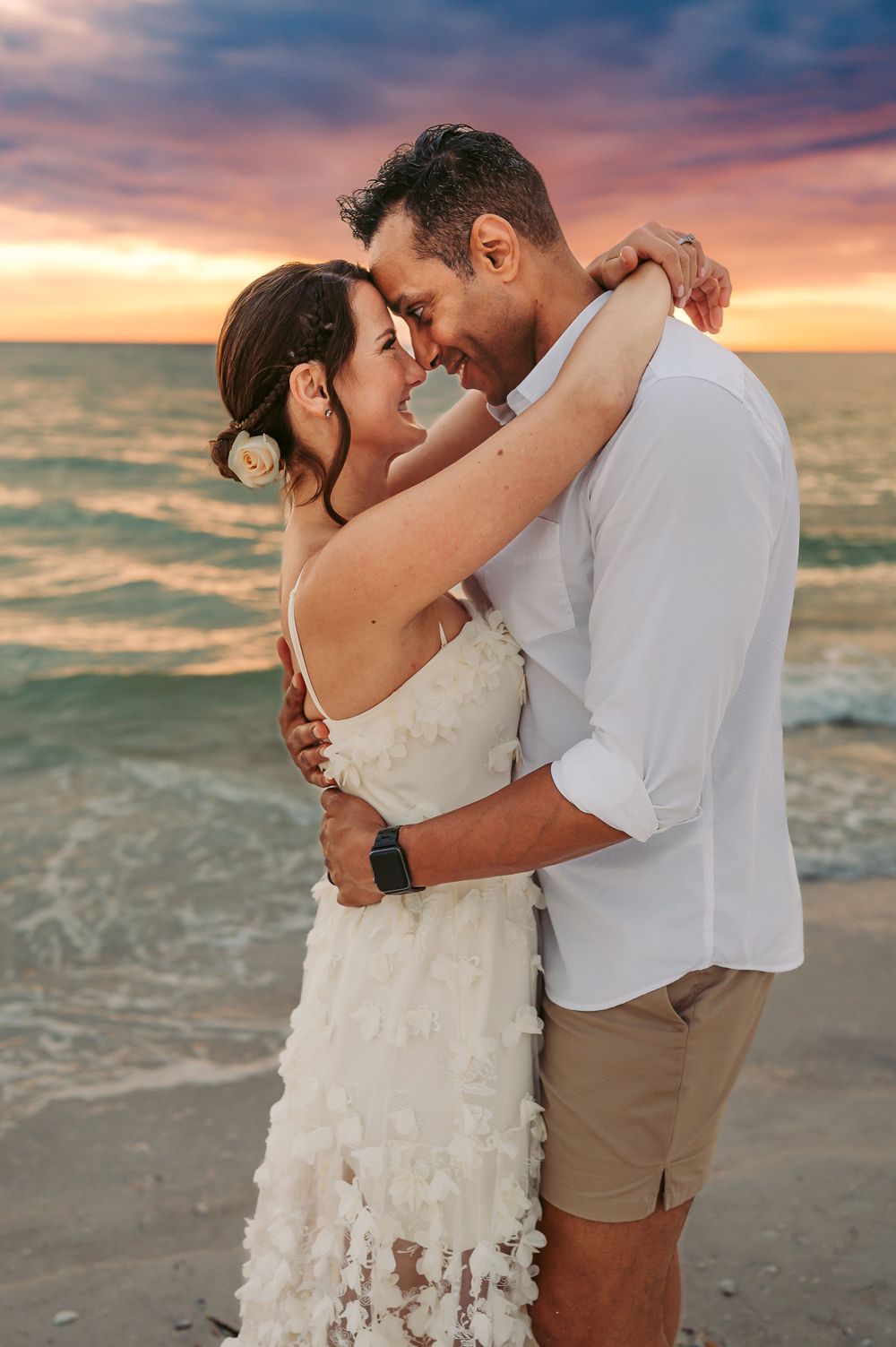 Woman standing with her arms wrapped around her husband's neck in front of the sunset at the beach in front of the beautiful Gulf coast.